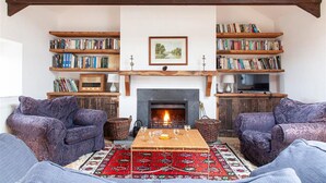 A cozy living room with a fireplace, two sofas, a coffee table, and bookshelves filled with books. - Carneggan Barn - Three Bedroom Cottage, Sleeps 6 (Lanteglos)