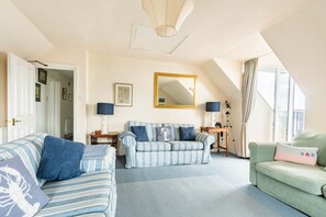 Un salon chaleureux avec un canapé à rayures, un fauteuil vert et un canapé bleu. On peut voir une table en bois avec une lampe, ainsi qu’un trépied sur lequel est posé un appareil photo.