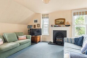 A cozy living room with a fireplace, two sofas, a television, and a bookshelf. - Bessborough Green - Two Bedroom Apartment, Sleeps 4 (St Mawes)