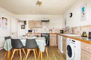 A compact kitchen with a dining area, a washing machine, and a clock on the wall. - Bessborough Green - Two Bedroom Apartment, Sleeps 4 (St Mawes)