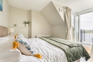 A bedroom with a bed, a window with curtains, a radiator, and a shelf with a plant. - Bessborough Green - Two Bedroom Apartment, Sleeps 4 (St Mawes)
