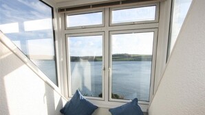 A corner window with a view of a body of water and a bench with cushions. - Bessborough Green - Two Bedroom Apartment, Sleeps 4 (St Mawes)
