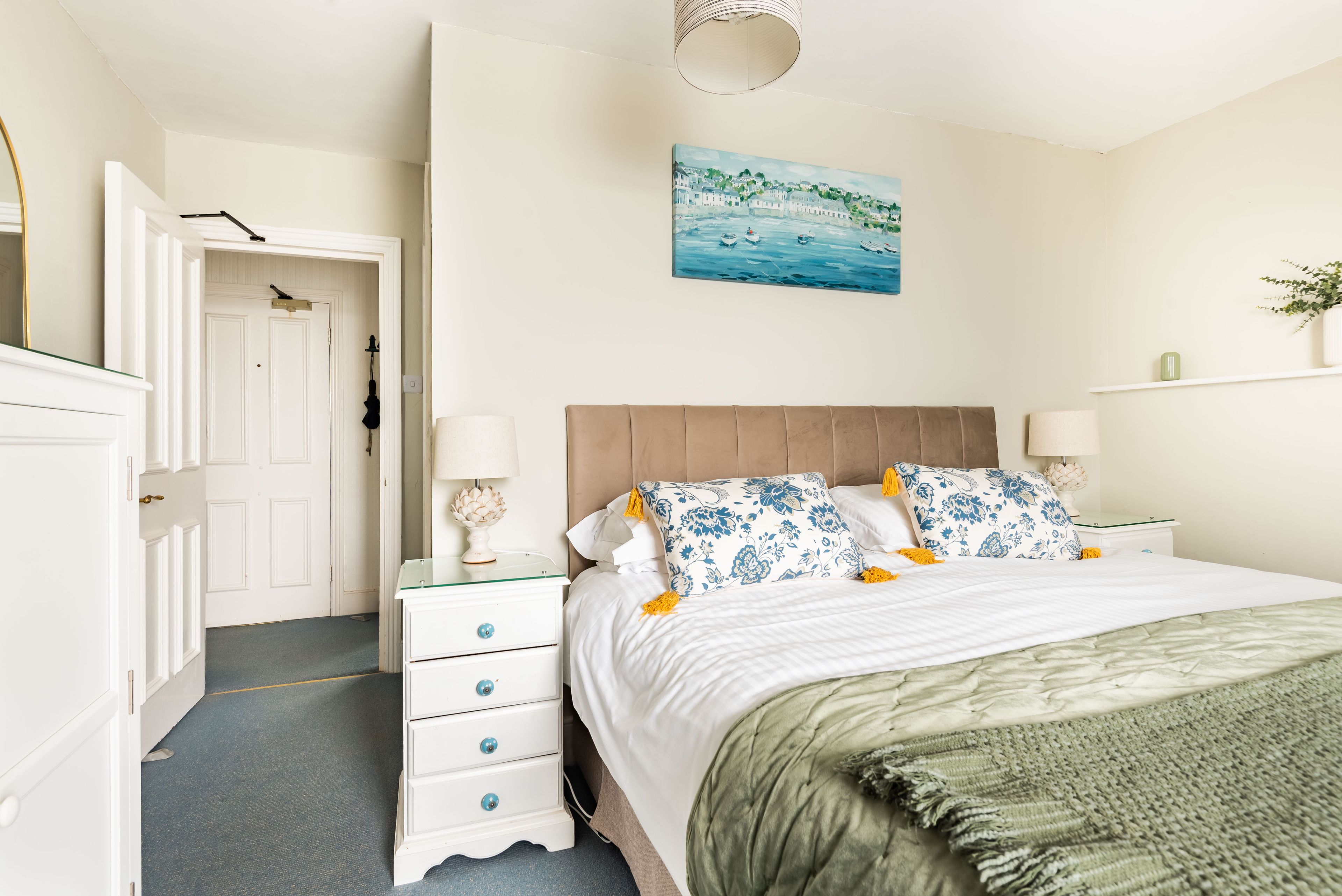 A bedroom with a bed, nightstands, a dresser, a painting, and a plant.