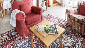 A spacious living area with a high ceiling, wooden beams, a staircase, a kitchen in the background, and a cozy seating area with a red sofa and patterned rug. - Lamellyon Roundhouse (Lanteglos)