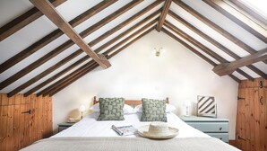 A bedroom with a sloped ceiling, wooden beams, a bed with patterned pillows, a nightstand with a lamp, and a decorative mirror. - Lamellyon Roundhouse (Lanteglos)