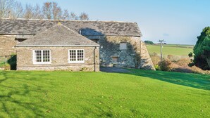 A stone house with a sloped roof, two windows with white frames, and a grassy lawn in front. - Lamellyon Roundhouse (Lanteglos)