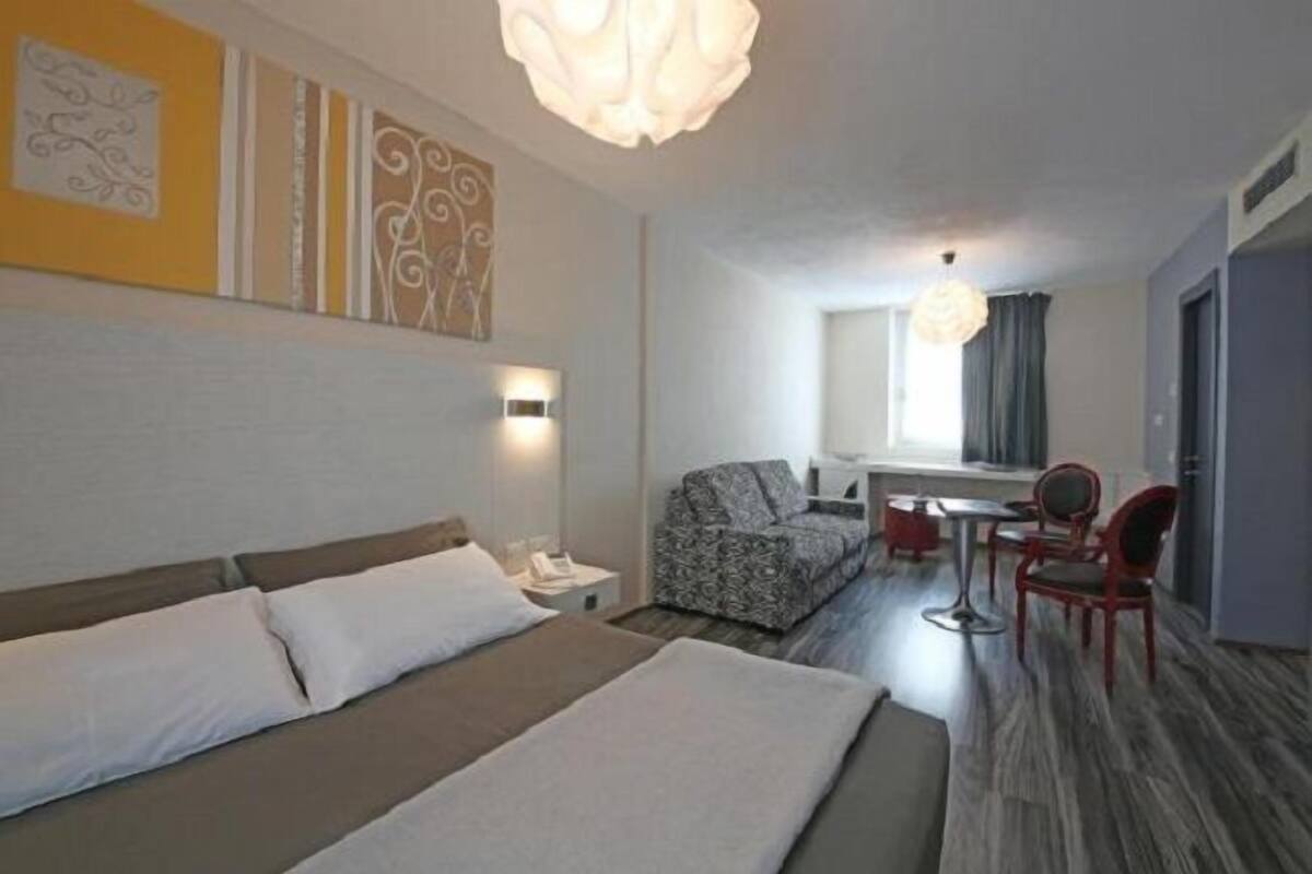 A hotel room with a large bed, a sofa, a small table, and chairs. There is a window with curtains and a ceiling light.