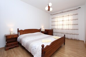 A bedroom with a wooden bed, two bedside tables, a nightstand, a window with curtains, and a hanging light fixture. - Apartment Robi (Rovinj)