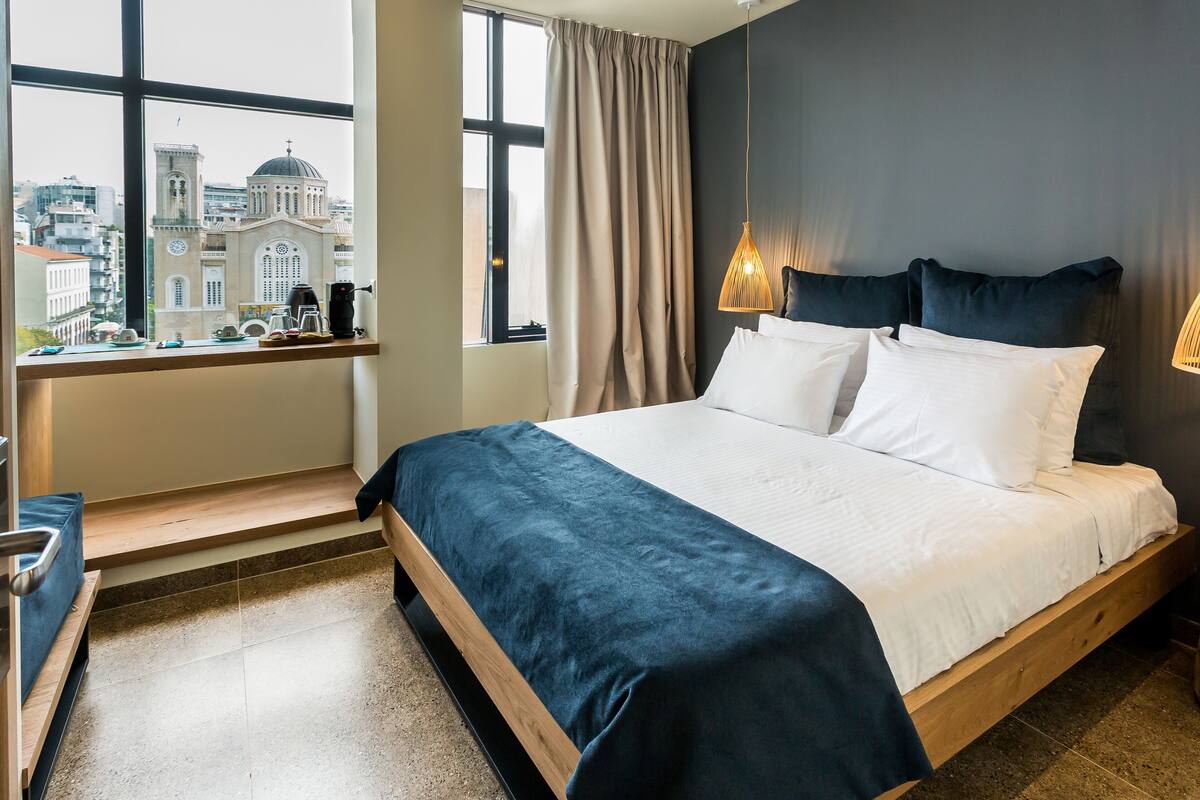 A hotel room with a large bed, a window with a city view, a desk with a lamp, and a chair.