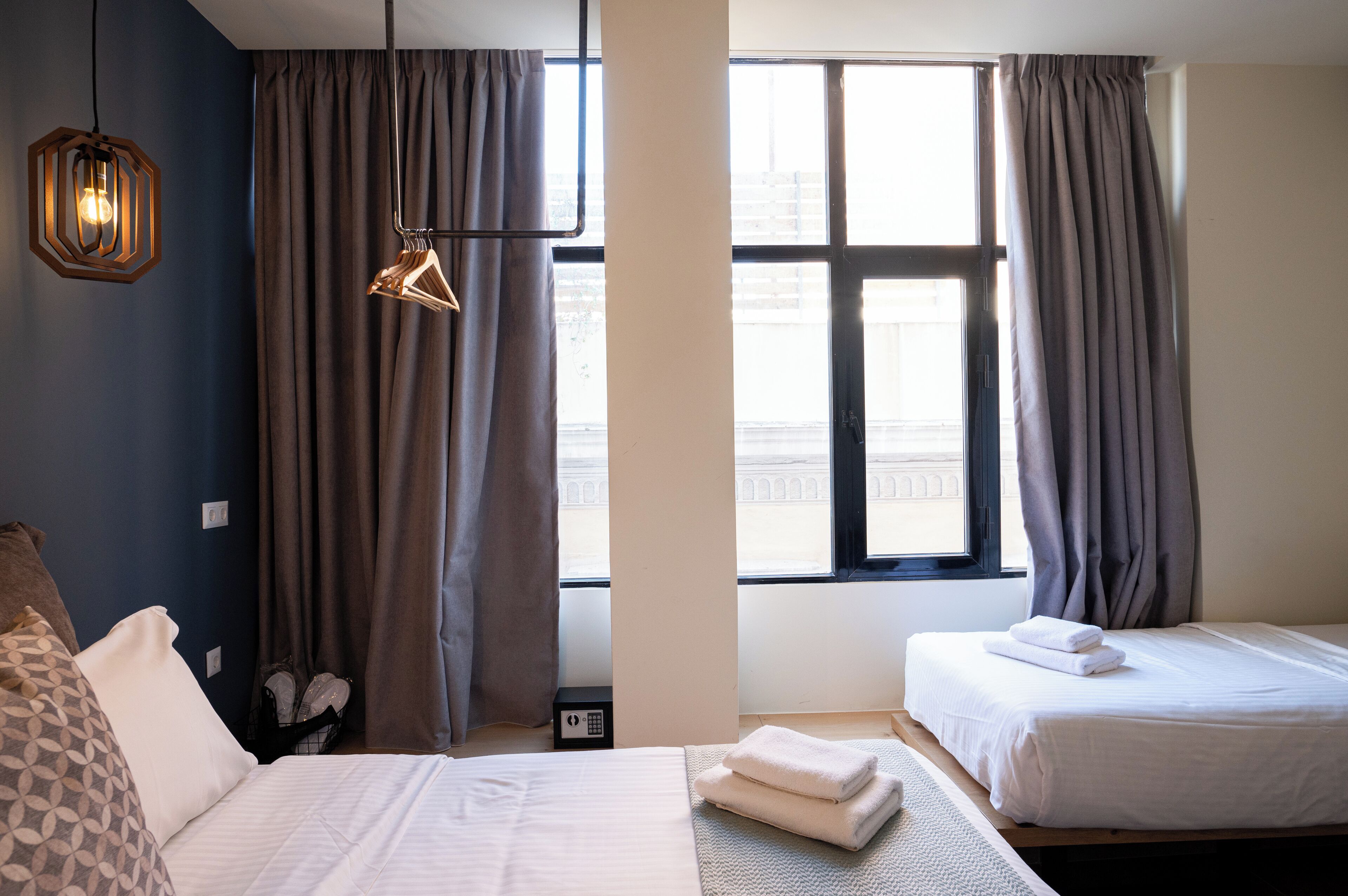 Une chambre d’hôtel avec deux lits, une grande fenêtre avec des rideaux, un luminaire suspendu et une table de chevet avec une lampe.