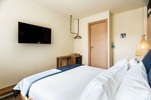 Quarto de hotel bem arrumado, com cama, escrivaninha, televisão, porta e luminária pendente.