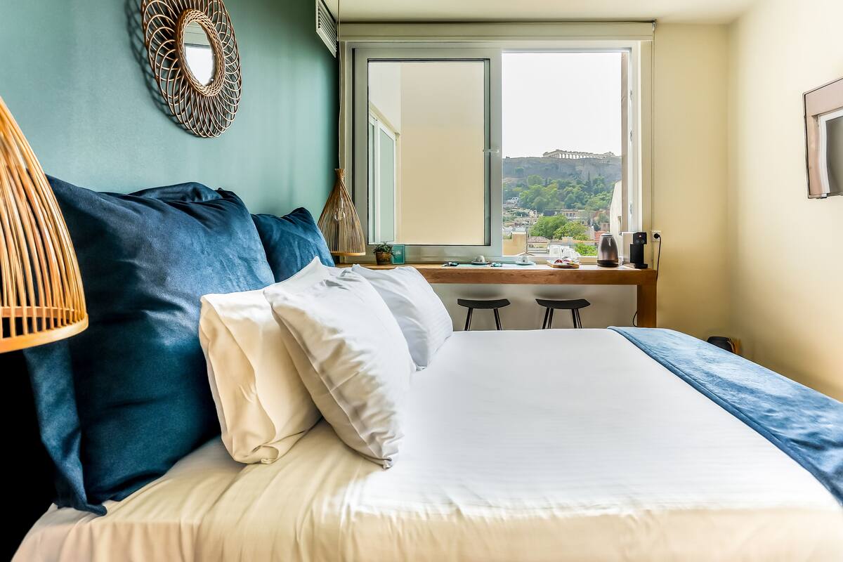 A neatly made bed with white and blue bedding, a wicker chair, and a view of a cityscape through a window.