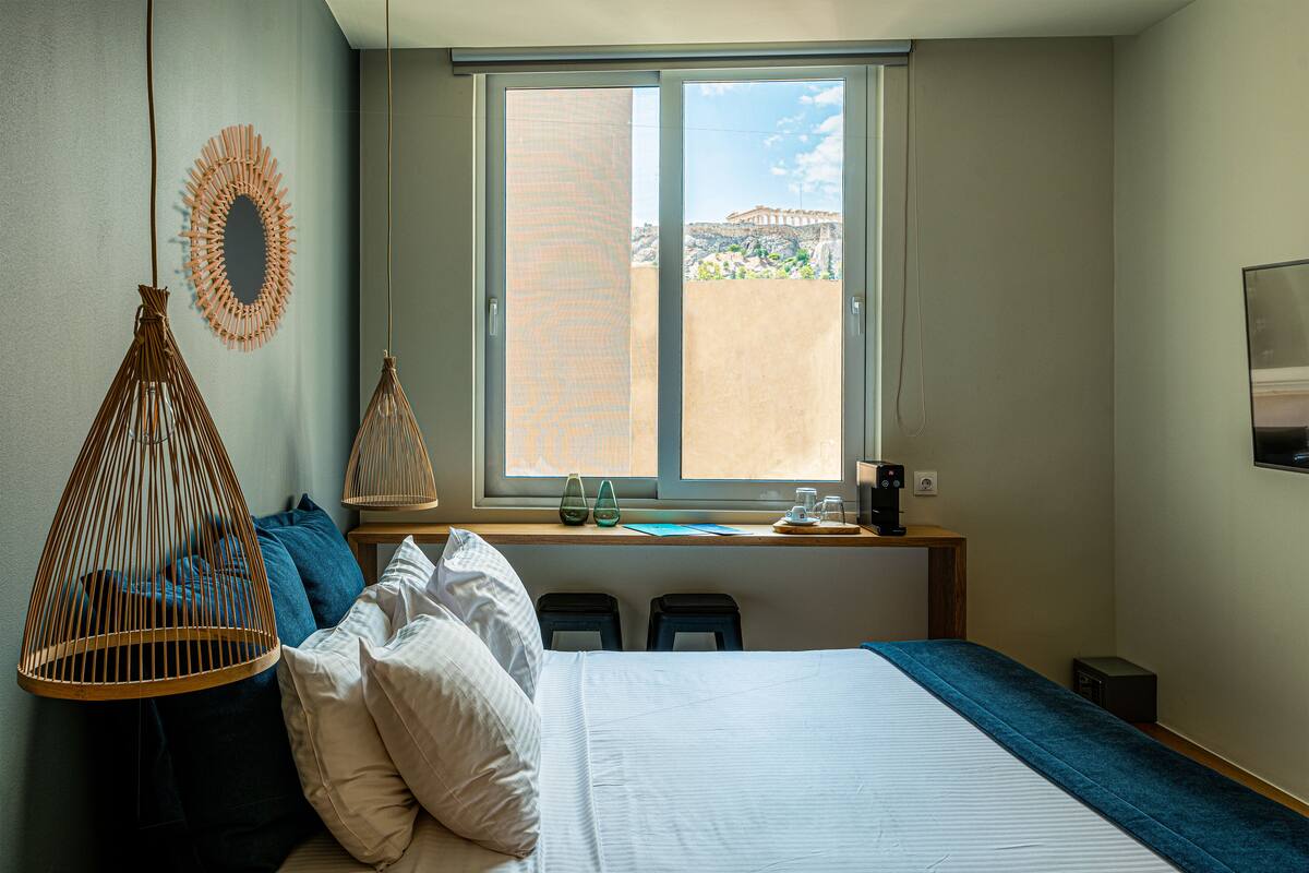 A bedroom with a bed, pillows, a hanging light fixture, a window with a view, and a desk with a coffee maker.