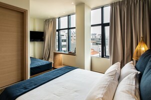 Quarto de hotel com uma cama grande, uma cama menor, uma TV fixada na parede e uma janela com cortinas.