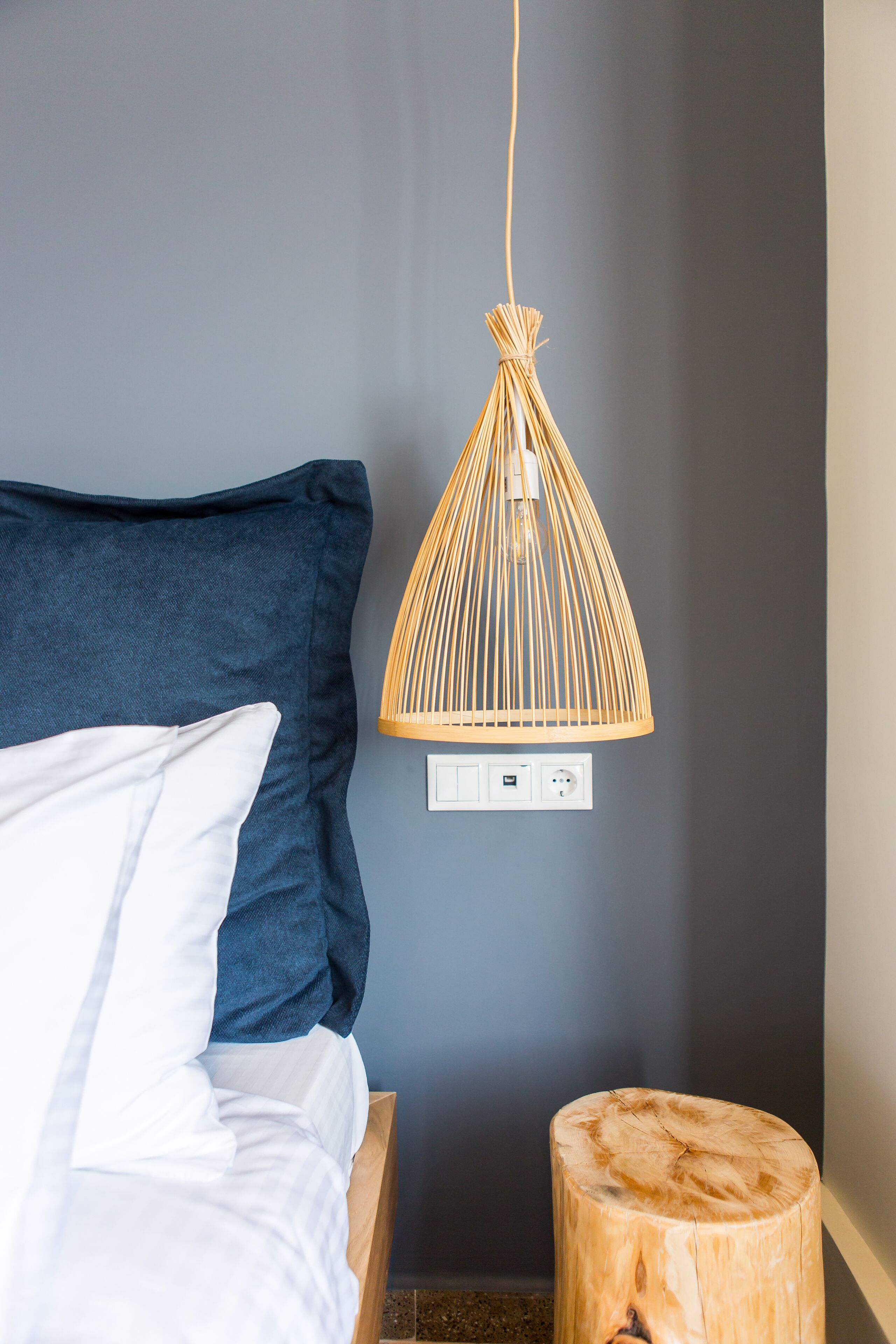 A bed with white and blue bedding, a wooden bedside table, and a woven pendant light fixture.