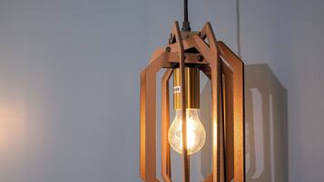 Luminária pendente hexagonal de madeira com lâmpada visível, suspensa por um fio preto em frente a uma parede lisa.