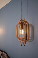 Luminária pendente hexagonal de madeira com lâmpada visível, suspensa por um fio preto em frente a uma parede lisa.