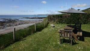 En udendørs siddeplads ved kysten med et bord og stole under et markise, med udsigt til en stenstrand og havet.