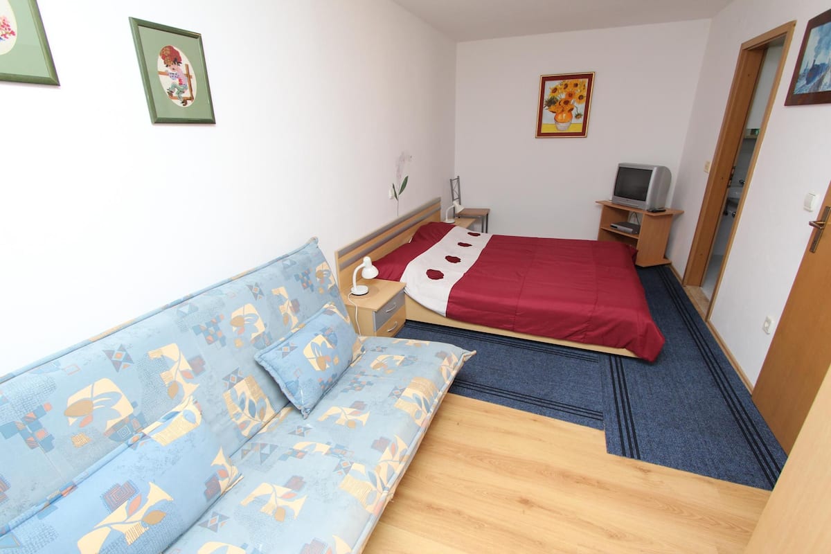 A small, well-lit room with a bed, a sofa, a TV, and a desk.