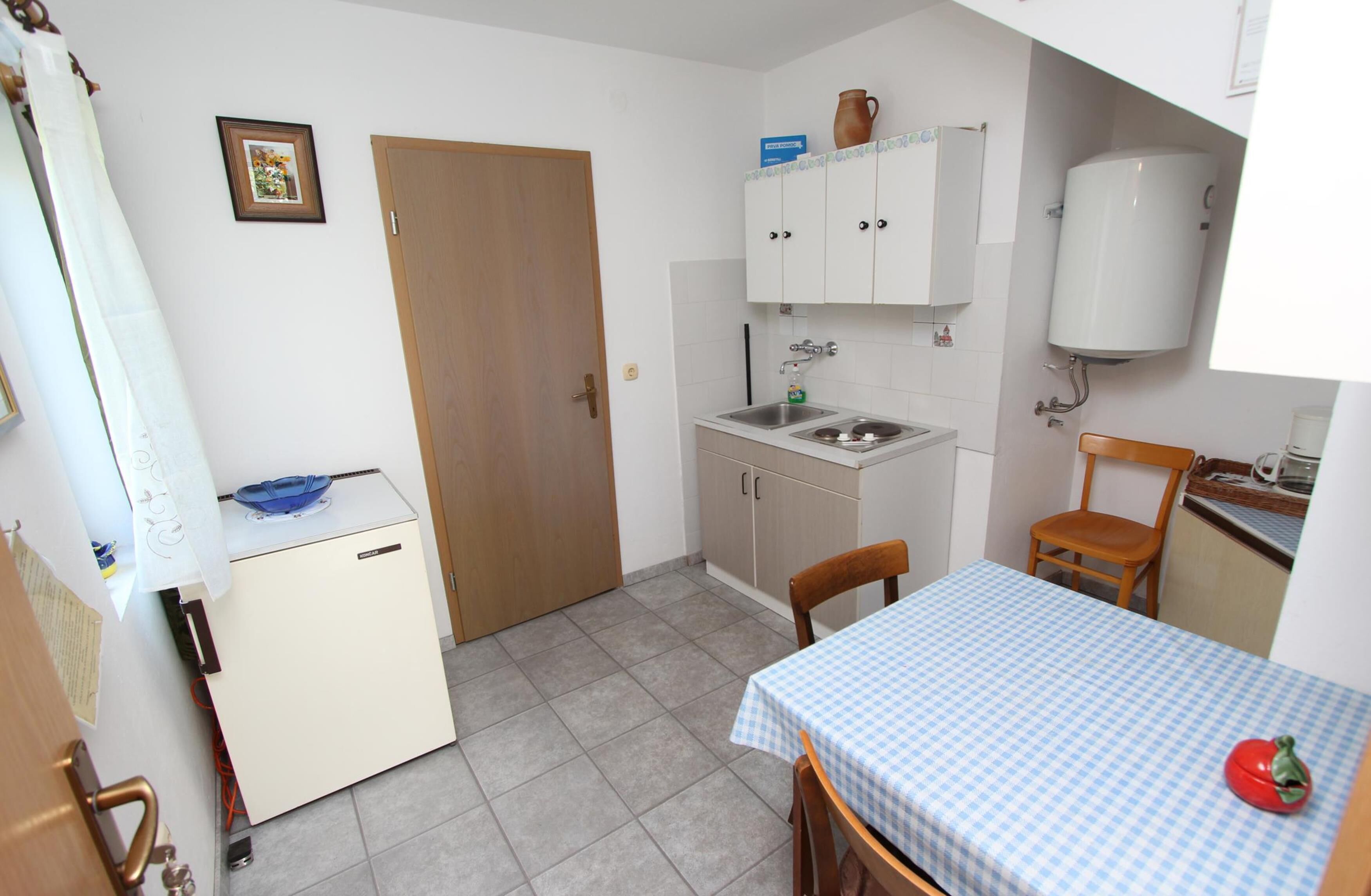 A compact kitchen with a refrigerator, sink, and cabinets. There is a small dining table with a checkered tablecloth and a chair. A wooden door and a picture frame are also visible.