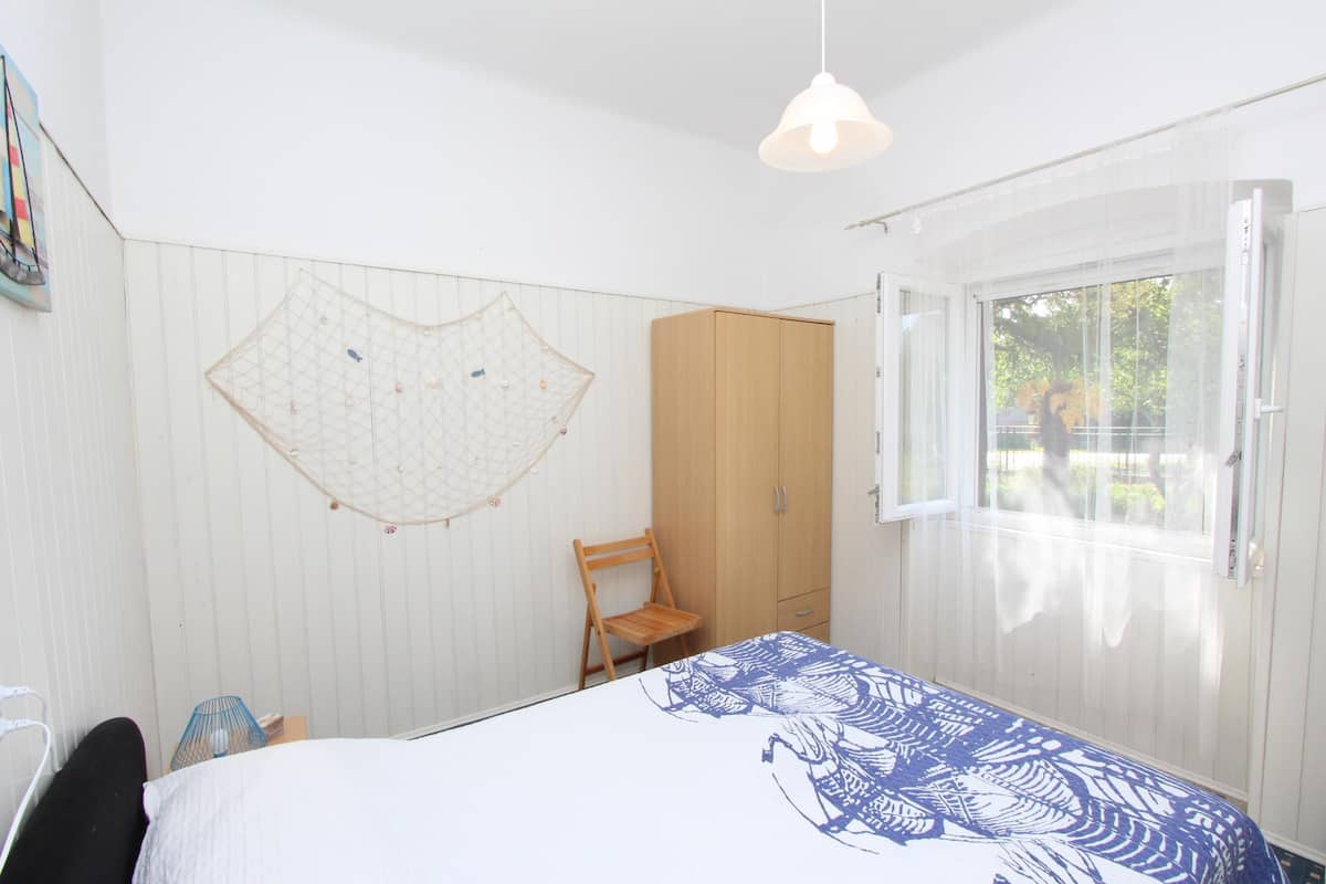 A bedroom with a bed, a wooden wardrobe, a chair, a hanging net, and a window with a view of the outside.