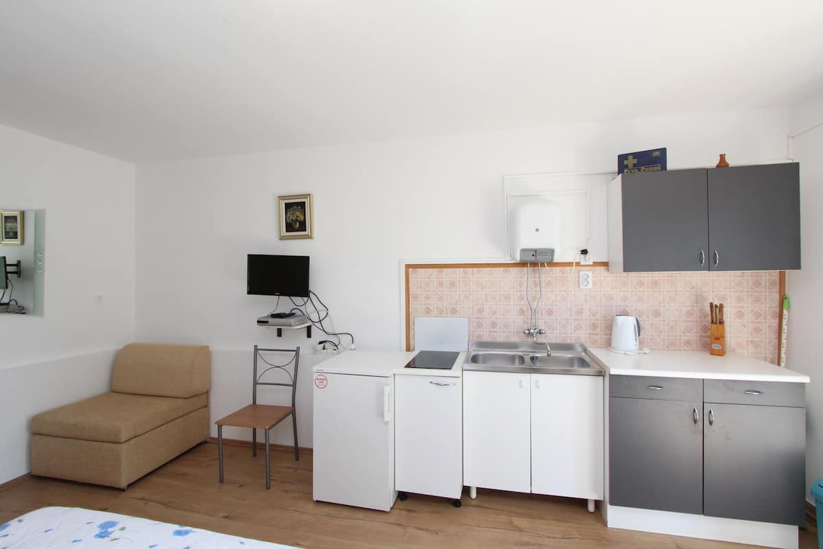 A compact room with a kitchenette, a small sofa, a chair, and a flat-screen TV.