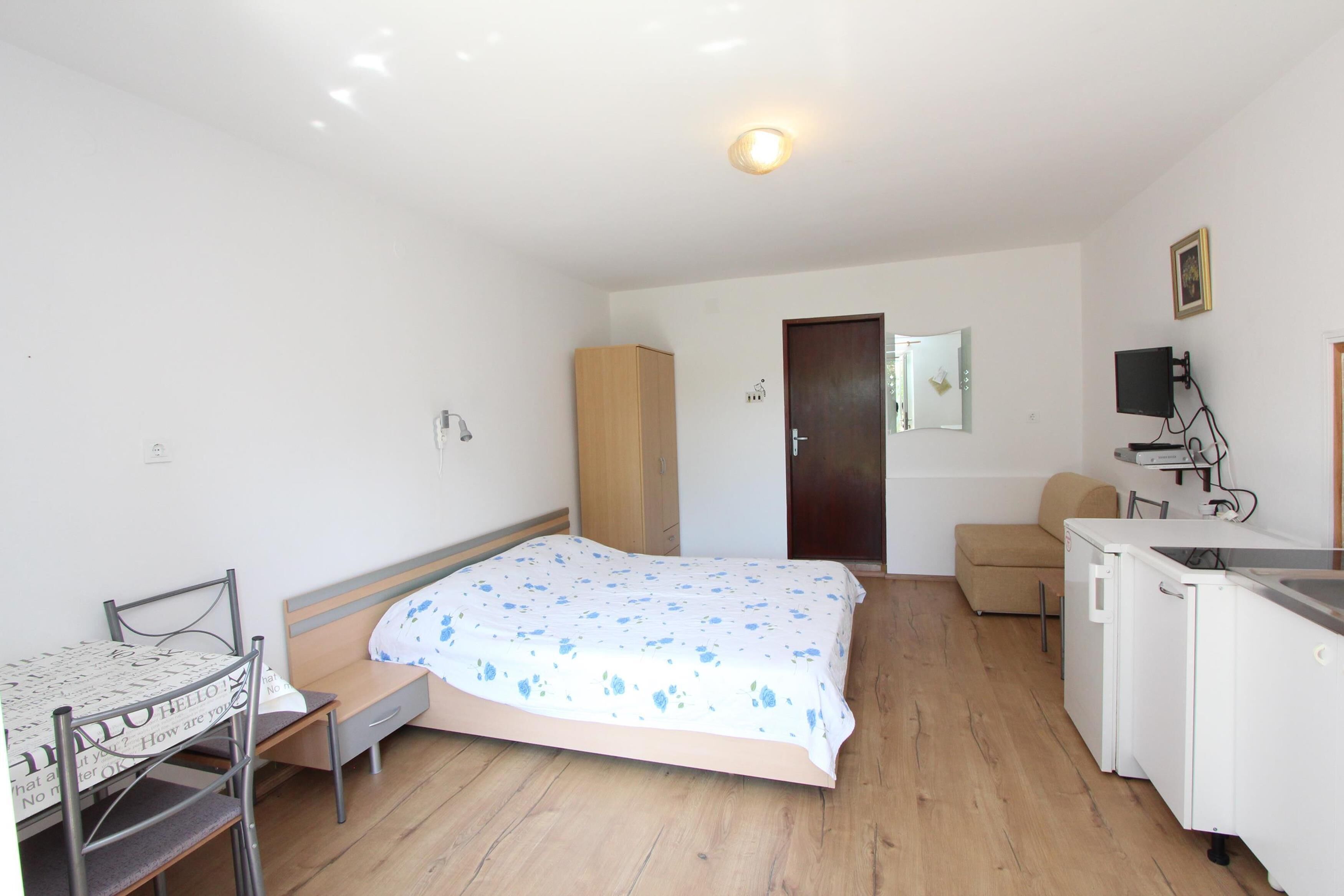 A small, well-lit room with a bed, a desk with a chair, a kitchenette, and a wardrobe.