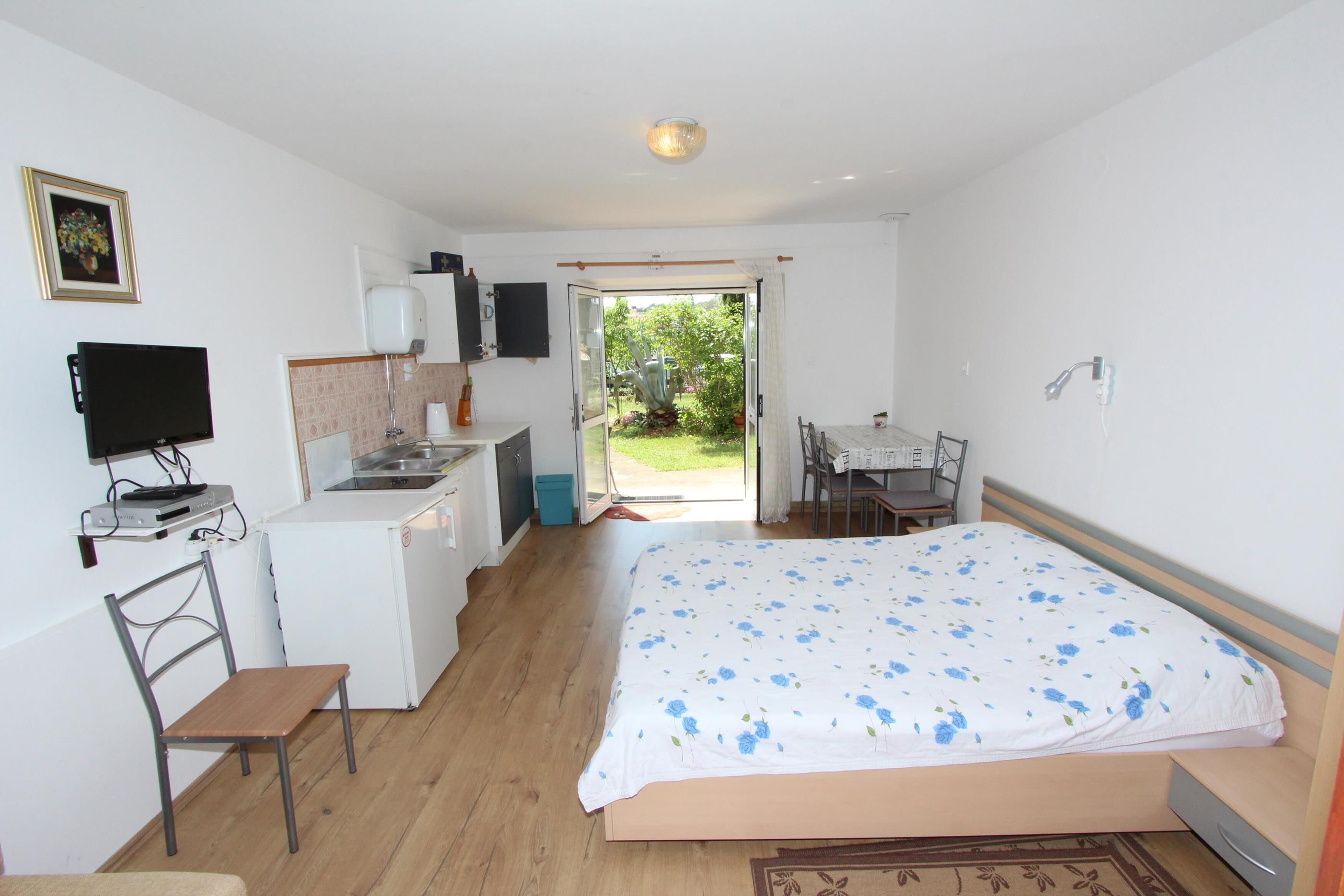Un dormitorio con cama, escritorio, silla, televisor, una pequeña kitchenette y una puerta corrediza de vidrio que da a un jardín.