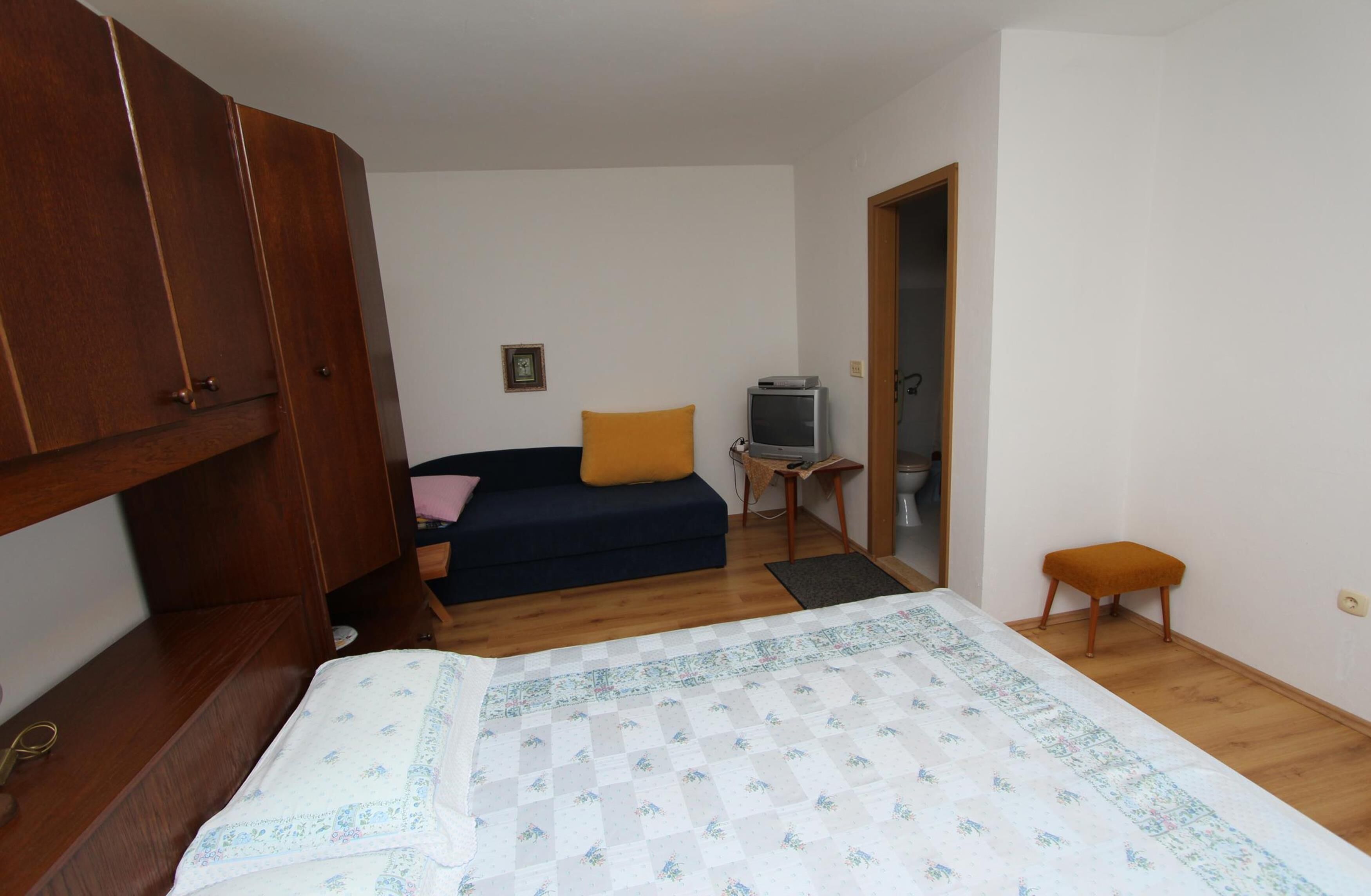A bedroom with a bed, a wooden wardrobe, a small table with a television, and a sofa with a yellow cushion.