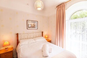 A neatly made bed with a headboard, two bedside lamps, a framed picture on the wall, and a window with curtains.