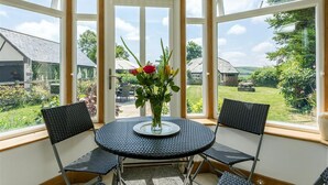 Une pièce d’angle avec une table ronde et deux chaises, un vase de fleurs et une vue sur un jardin et des bâtiments à l’extérieur.