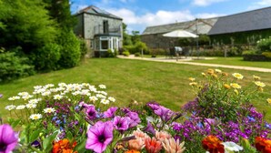 Un jardin où s’épanouit une grande variété de fleurs au premier plan, un bâtiment en pierre en arrière-plan et un ciel bleu dégagé.
