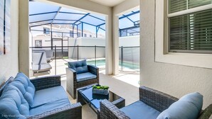 A covered patio area with wicker furniture, a glass table, and a view of neighboring buildings. - Champions Gate Exec Villas- 1571sand (Davenport)