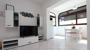 A modern living room with a flat-screen TV, white entertainment unit, dining table with chairs, and a window with a view. - Apartments Rozi (Rovinj)