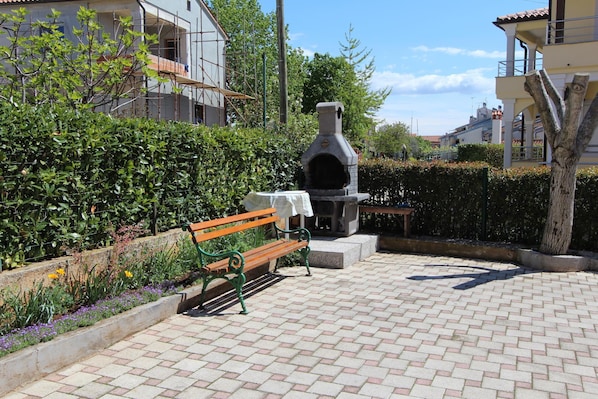 Relax on the patio with a brick floor, featuring a bench and table, surrounded by lush hedges and flowering plants.
