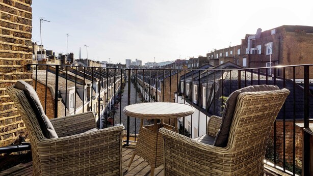 A balcony with wicker furniture and a table, overlooking a cityscape with buildings and a clear sky.