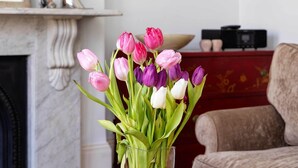 A vase with pink and white tulips on a wooden table in a room with a fireplace, a framed picture on the wall, and a red cabinet.