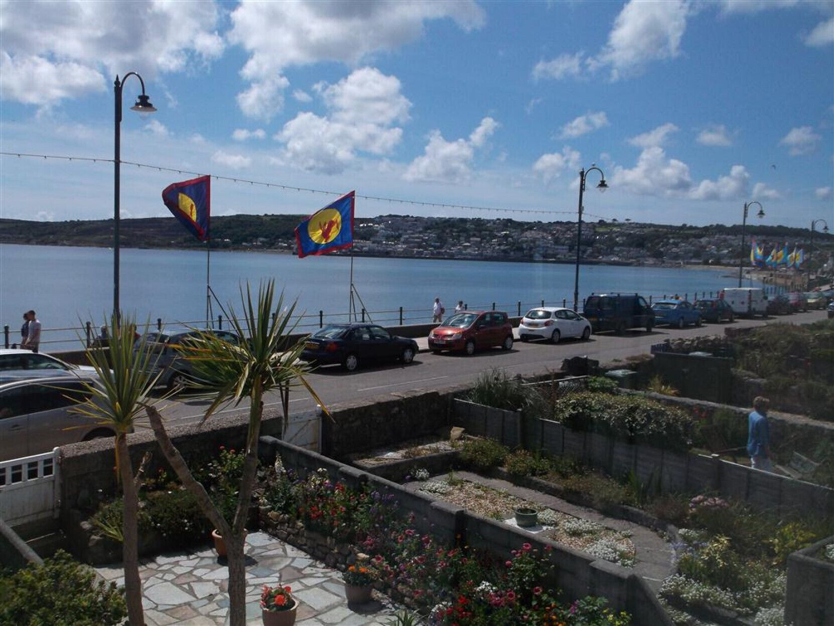 A coastal road with parked cars, a flag, and a view of the sea and town in the background.