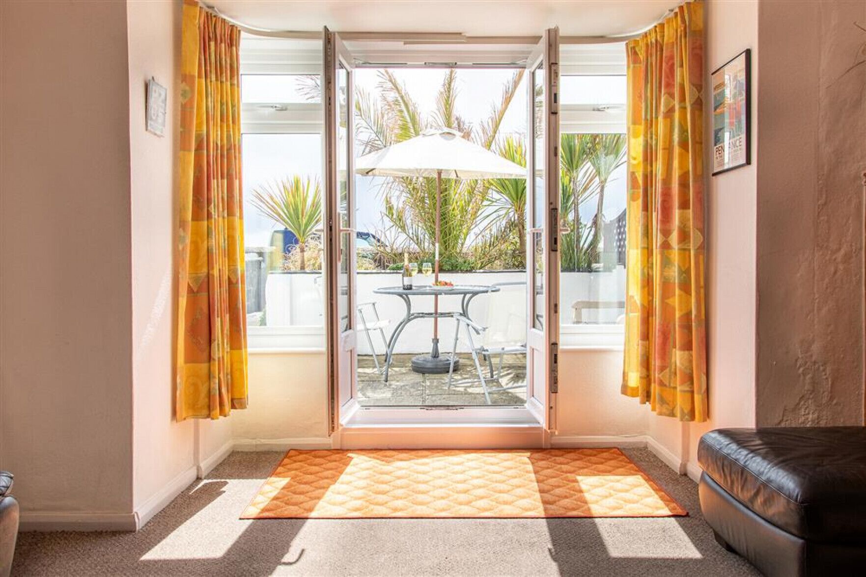 Quarto com porta de vidro deslizante que dá para área externa com mesa e cadeiras, sofá de couro preto e tapete estampado.