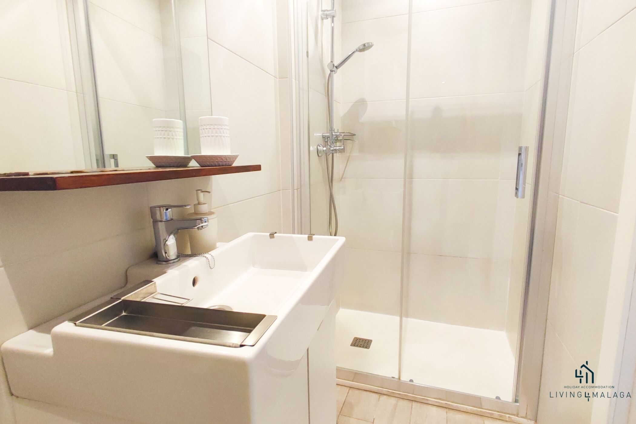 Une salle de bain moderne avec une douche à parois de verre, un lavabo blanc doté d’un distributeur de savon et une étagère en bois au-dessus du lavabo.