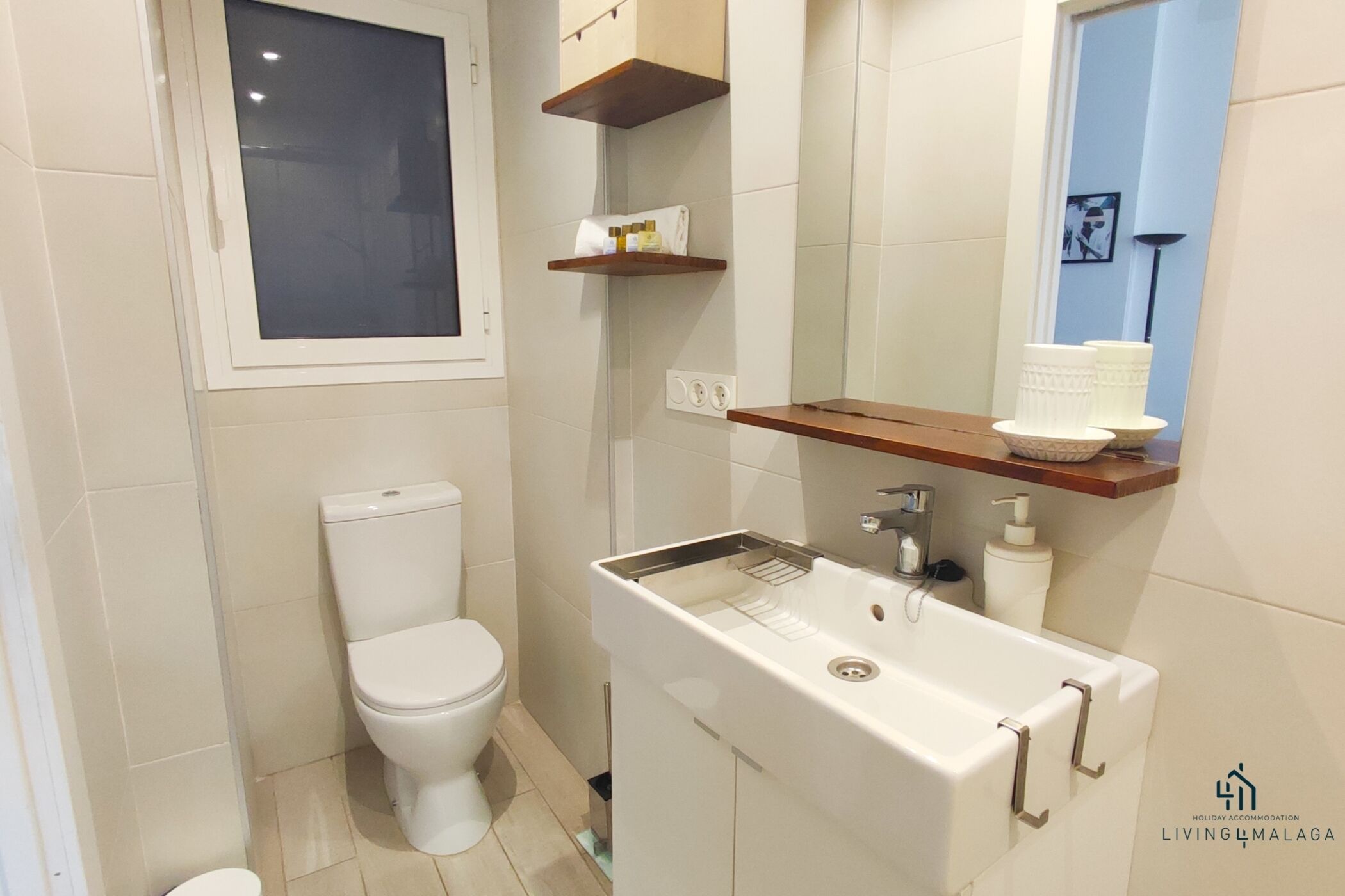 Un bagno con un water, un lavandino e uno specchio. Ci sono scaffali sopra il lavandino e una finestra sopra il water.