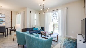 A modern living room with a sofa, two armchairs, a coffee table, and a TV. There is a dining area with a table and chairs, and large windows with curtains. - Champions Gate Exec Villas- 8983sd (Davenport)