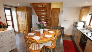 A dining area with a wooden table set for four, surrounded by wooden chairs. A vase with flowers is on the table, and there is a staircase leading to another room. - Skylark - Three Bedroom Cottage, Sleeps 6 (Camborne)