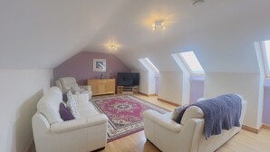 A loft bedroom with a sloped ceiling, a TV, a sofa, a chair, a cabinet, a rug, and skylights. - Skylark - Three Bedroom Cottage, Sleeps 6 (Camborne)