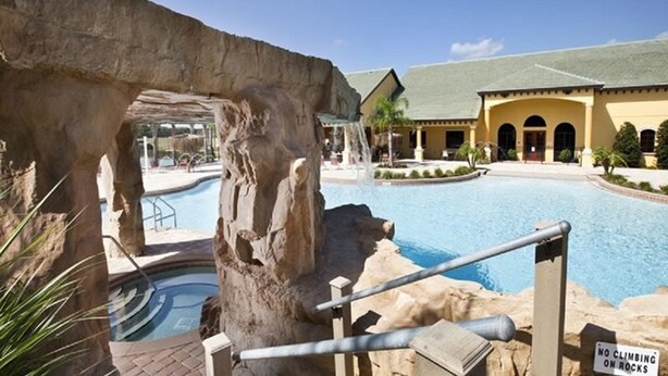 Un área de piscina con un arco natural de roca, un cartel que prohíbe subir a las rocas y un edificio con techo verde al fondo.