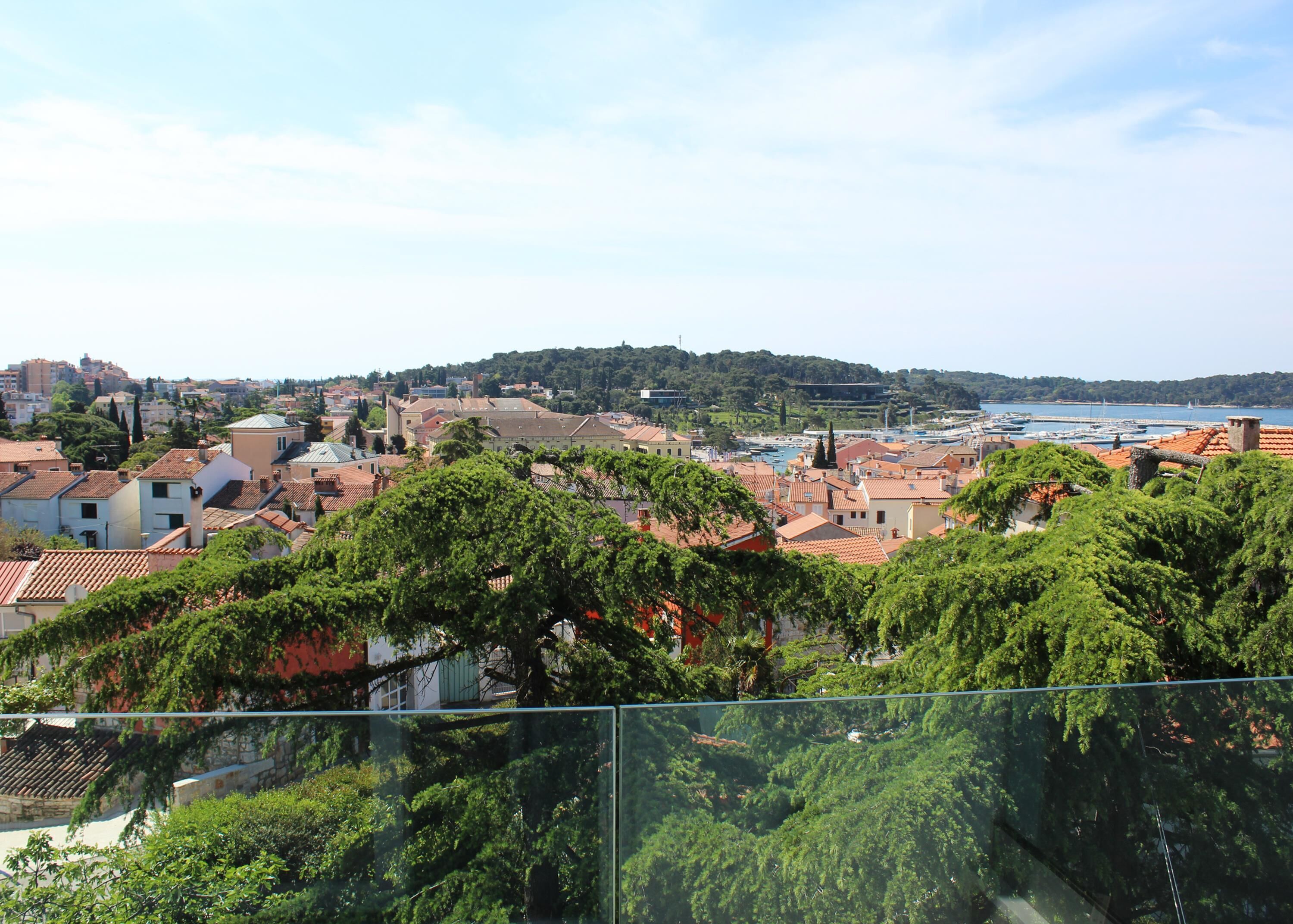 A coastal town with terracotta-roofed buildings, a marina, and a hill with greenery in the background.