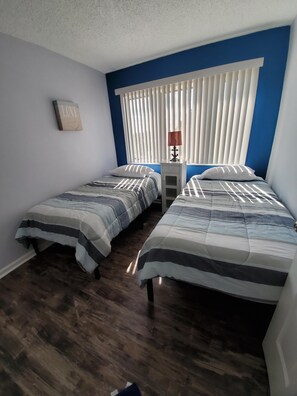 Two single beds with striped bedspreads, a table with a lamp, and a framed picture adorn this cozy space.