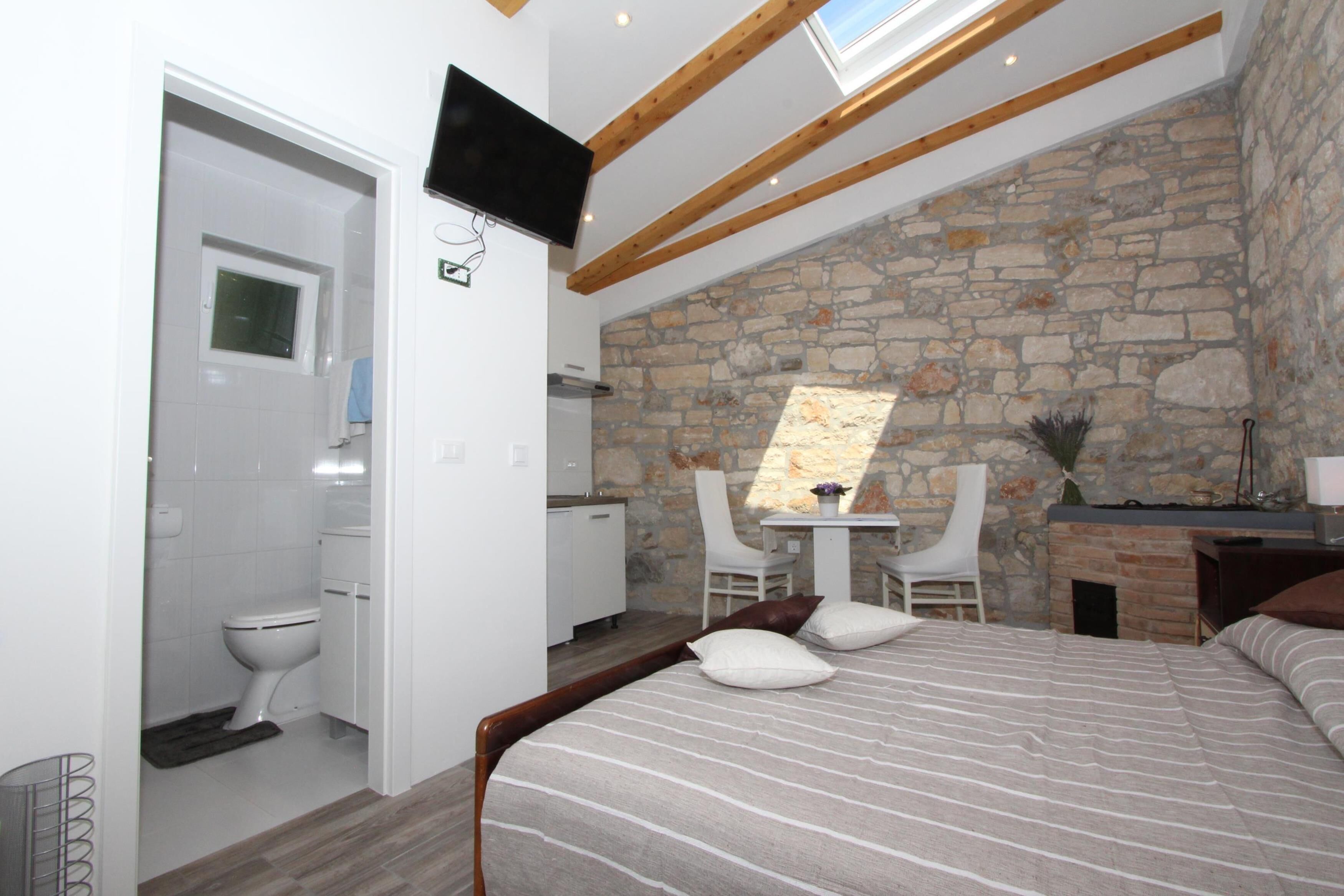 A bedroom with a stone wall, a bed with striped bedding, a desk with a chair, and a TV mounted on the wall.