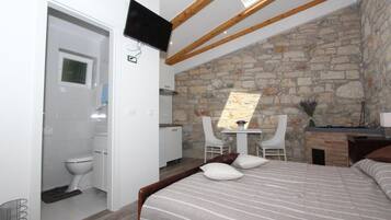 A bedroom with a stone wall, a bed with striped bedding, a desk with a chair, and a TV mounted on the wall.