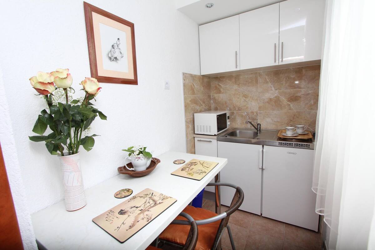 Une petite cuisine équipée avec des meubles blancs, un four à micro-ondes et un évier. On y trouve une table ornée d’un vase de fleurs et de quelques assiettes décoratives.